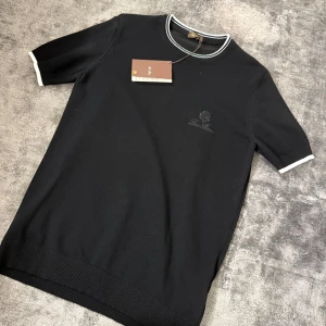Loro Piana T-shirt - Svart t-shirt från Loro Piana med diskret broderad logga på bröstet. Rund hals med vit och svart ribbad kant, samt vita detaljer vid ärmslut. Klassisk och stilren design i mjuk bomull som känns lyxig mot huden. Perfekt för dig som gillar minimalistisk stil.