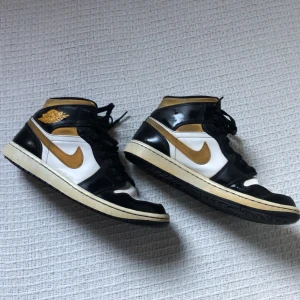 Nike Air Jordan 1 svart/vit/guld - Säljer ett par Nike Air Jordan 1 med svart, vit och guld färgkombination. Klassisk high-top siluett med svarta snören, guldfärgade detaljer och logga på sidan. Ovandel i skinn och textil, perforerad tå och ikonisk Jordan-vinge på ankeln. Perfekta för dig som gillar streetstyle.
