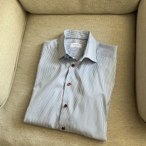 Randig skjorta från Eton - Snygg blå- och vitrandig skjorta från Eton i klassisk modell. Skjortan har röda knappar som ger en cool kontrast och är tillverkad i mjuk bomull. Perfekt för dig som gillar stilrena och fräscha plagg med en twist. Använd en gång .