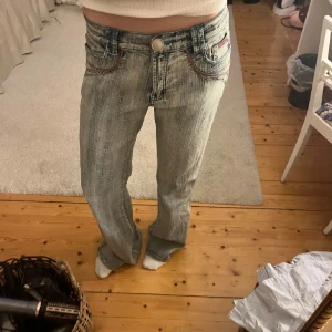 Grå bootcut jeans med broderi - Snygga jeans med unika broderade detaljer baktill. 💕De har en lågmidjad passform och en bootcut stil. Jeansen har också blåa samt röd/rosa sömmar. Ytterbensmått 109cm Innerbensmått 86 cm Midjemått 41 cm rakt över