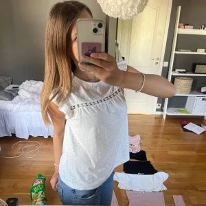 Vit blus med spetsdetaljer - Säljer en söt vit blus med broderade hålmönster och spetsdetalj längs bröstet. Blusen har korta volangärmar och rund hals. Perfekt till jeans eller kjol för en fräsch look. Materialet känns lätt och luftigt, passar dig som gillar romantisk stil.