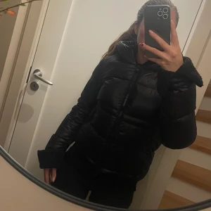 Svart pufferjacka  - Säljer en svart pufferjacka från refrigiwear stor huva och dragkedja framtill. Jackan har en snygg badge på ärmen och är vadderad för extra värme. Perfekt för kalla dagar dock är det en kort jacka som slutar lite innan höften. Kan mötas upp inom Stockholms området