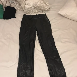 Svarta skinnbyxor från New York  - Säljer ett par svarta skinnbyxor från New Yorker  i storlek 34. Byxorna har en slim passform, dragkedja i sidan och elastisk midja för extra komfort. Snyggt glansigt material som ger en edgy look. Perfekta för dig som vill sticka ut lite extra.