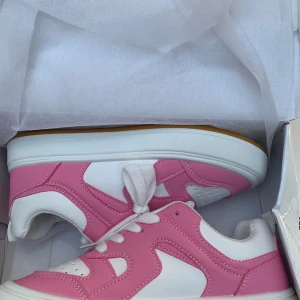 Vita och rosa sneakers med snörning - Snygga sneakers i vitt och rosa med klassisk design och tjock vit sula. Skorna har perforerade detaljer framtill och breda vita skosnören. Perfekta för dig som vill sticka ut med färg och stil. Tillverkade i syntetmaterial.