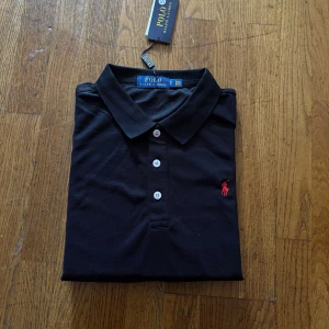Svart Polo Ralph Lauren t-shirt S - Svart t-shirt från Polo Ralph Lauren med klassisk pikékrage och tre vita knappar framtill. Röd broderad logga på bröstet ger en clean och stilren vibe. Materialet är mjuk bomull som känns skönt mot huden. Perfekt för dig som gillar enkel och snygg design.