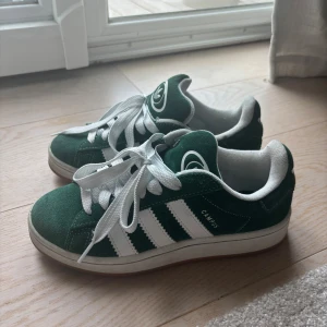 Adidas Campus sneakers gröna mocka - Gröna Adidas Campus sneakers i mocka med klassiska vita ränder och vit logga på häl och plös. Skorna har vit insida, vita skosnören och en snygg gummisula i brunt. Perfekt retrostil med låg profil och rund tå. 