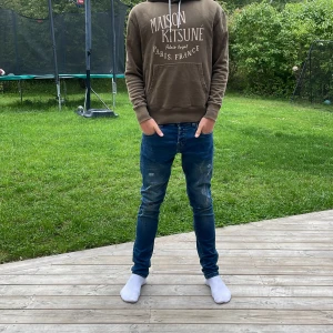 Maison Kitsuné grön hoodie med tryck - Säljer en grön hoodie från Maison Kitsuné med vit text på bröstet. Tröjan har huva med vita snören och en stor magficka. Perfekt för dig som gillar streetwear och vill ha något snyggt och bekvämt.