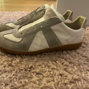 Maison Margiela Gats - !Intressekoll!Maison Margiela sneakers med vit läderbas och grå mockadetaljer. Klassisk gummisula i brunt och rund tå. Skorna har snörning och kommer med dustbags och originalkartong. Perfekta för dig som gillar stilrena och lyxiga sneakers.