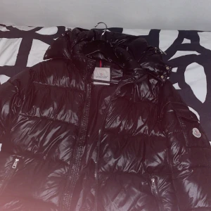 Svart Moncler pufferjacka med huva - Säljer en svart pufferjacka från Moncler med glansig finish och huva. Jackan har dragkedja framtill, fickor med dragkedja och en extra ficka på ärmen med Moncler-logga. Perfekt för kalla dagar och riktigt snygg streetstil. Och märket funnets och scanna men kedjan har gått av fast det är nåt som går och fixa jätte enkelt och därför säljer jag den så billigt 