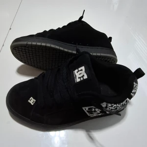 Svarta DC sneakers med grafiska detaljer - Svarta sneakers från DC Shoes med bred plös, snörning och coola vita grafiska tryck på hälen. Skorna har en platt sula och rund tå, tillverkade i mocka för en skön och streetig känsla. Perfekta för dig som gillar skate-stil och Grisch funkar också för orten😆