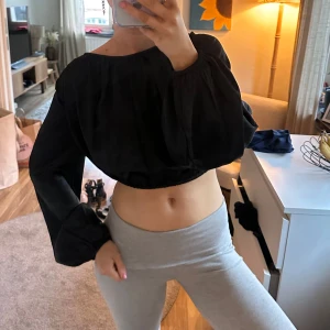 Svart croppad blus med knyt i ryggen - Säljer en svart croppad blus med vida långa ärmar och snygg knytning i ryggen som går att justera. Det är en kort blus! Går över naveln😌typ glansig material. Från NA-KD i XS