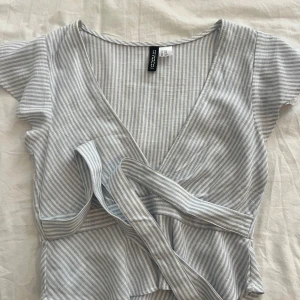 Randig omlottopp från H&M Divided - Säljer en ljusblå och vit randig omlottopp från H&M Divided. Aldrig använd men den va tyvär förliten på mig.