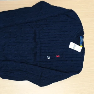 Marinblå stickad tröja Polo Ralph Lauren - Säljer en marinblå stickad tröja från Polo Ralph Lauren med klassiskt kabelstickat mönster och röd broderad logga på bröstet. Tröjan har rund hals och långa ärmar, perfekt för dig som gillar stilren och tidlös design.