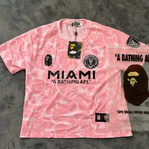 Rosa BAPE x Miami kortärmad t-shirt - Snygg rosa t-shirt från BAPE x Miami med camomönster, stora tryck fram och bak samt klassisk BAPE-logga. Kortärmad modell i mjuk bomull, perfekt för dig som vill sticka ut med streetwear-vibe. Nummer 93 och BAPE-tryck på ryggen.