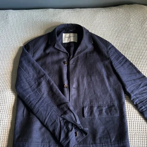 Mörkblå overshirt från These Glory Days - Snygg mörkblå overshirt från These Glory Days i linne. Jackan har klassisk krage, knäppning med tre knappar och två stora fickor framtill. Perfekt lager-på-lager-plagg med stilren och minimalistisk look.