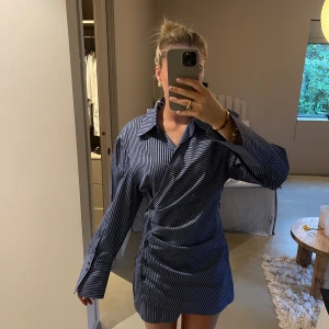 Randig skjortklänning från & Other Stories - Mörkblå kort skjortklänning från & Other Stories med vita ränder, lång ärm och knappar längs sidan. Klänningen har klassisk krage, markerade manschetter och en snygg drapering över höften.