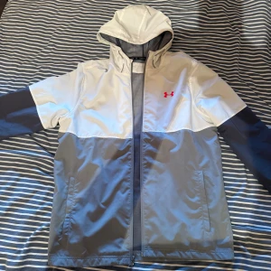 Grå och vit vindjacka från Under Armour - Snygg vindjacka från Under Armour i grått och vitt med röd logga på bröstet. Jackan har huva, hel dragkedja och två fickor framtill. Tillverkad i polyester och har meshfoder på insidan för extra komfort. Perfekt för blåsiga dagar.