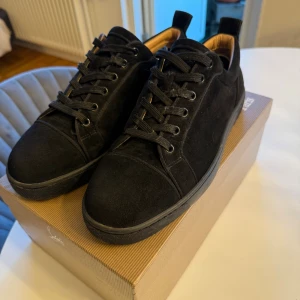 Svarta mockasneakers från Louboutin - Stilrena svarta sneakers från Christian Louboutin i mjuk mocka med svarta snören och diskret rund tå, storlek 43. Snygg och minimalistisk design med svart sula och lyxig känsla. Perfekta för dig som gillar exklusiva och tidlösa skor.