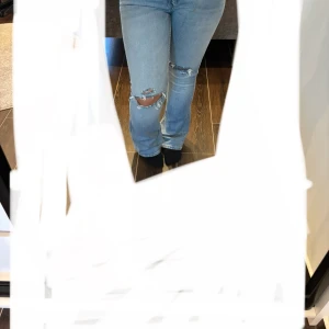 Ljusblå slitna jeans med bootcut - Säljer ett par ljusblå jeans med slitningar och hål på knäna. Jeansen har klassisk femficksdesign, bootcut-ben och är tillverkade i mjukt bomullsjeans. Perfekta för en avslappnad och trendig look.