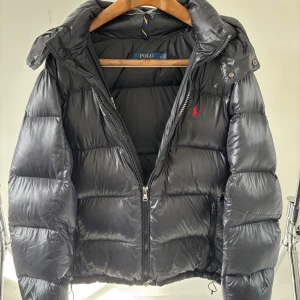 Svart dunjacka från Polo Ralph Lauren - Svart, glansig dunjacka från Polo Ralph Lauren med röd broderad logga på bröstet. Jackan har huva, dragkedja framtill och flera fickor med dragkedja. Puffig och varm modell som är perfekt för kalla dagar. Klassisk och stilren look. Köpt på Care of Carl för 6700 för 2 vintrar sedan och väl behållen sedan dess. Pris kan diskuteras.
