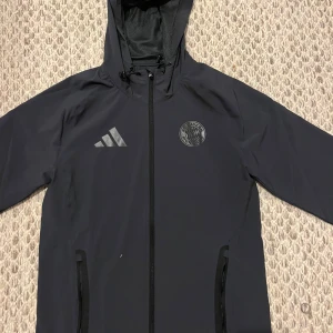 Svart vindjacka från Adidas x Bayern - Svart vindjacka med huva från Adidas i samarbete med FC Bayern München. Jackan har dragkedja framtill, två fickor med dragkedja och klubbens logga på bröstet. Perfekt för dig som gillar sportig stil och vill representera ditt lag.