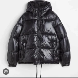 Svart pufferjacka med huva - Säljer en svart, glansig pufferjacka med huva och dragsko nertill. Jackan har dragkedja framtill och två fickor på sidorna. Perfekt för kalla dagar och ger en riktigt chill vibe. Passformen är oversized och jackan är riktigt mjuk och varm.