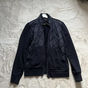 Mörkblå cardigan Tommy Hilfiger S - Mörkblå cardigan från Tommy Hilfiger i storlek S. Jackan har quiltade partier framtill, hög krage och dragkedja. Tillverkad i polyester med mjuka muddar vid ärmslut och midja. Snygg och stilren med diskret logga på ärmen.