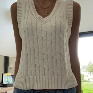 Vit kabelstickad väst med v-ringning - Säljer en vit kabelstickad väst med bred ribbad kant och djup v-ringning. Västen är ärmlös och har en klassisk, lite croppad passform som funkar perfekt över en skjorta eller t-shirt. Enkel att styla och superfräsch till jeans.