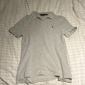 Grå pikétröja Polo Ralph Lauren XL (barnstorlek) - Snygg ljusgrå pikétröja från Polo Ralph Lauren i slim fit-modell. Klassisk krage, korta ärmar och knäppning med två knappar framtill. Broderad logga på bröstet. Perfekt till jeans eller shorts för en clean look. (Jag är 175cm lång och den passar perfekt på mig)
