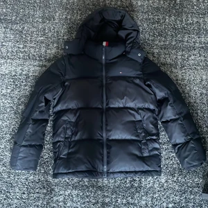 Tommy Hilfiger vinterjacka  - Säljer en svart pufferjacka från Tommy Hilfiger med huva och dragkedja. Jackan har en clean look med diskret logga på bröstet och snygga detaljer på insidan. Perfekt för kalla dagar och riktigt skön att bära. Kan slipa lite på priset vid snabb affär.