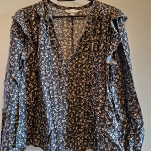 Blommig blus med volang från H&M - Supersnygg blommig blus från H&M i mörkblått med vita blommor. Blusen har v-ringning, långa ärmar och volangdetaljer på axlarna. Perfekt för dig som gillar romantiska och trendiga plagg med lite extra detaljer.