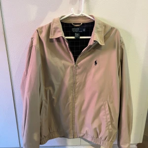 Beige vindjacka från Polo Ralph Lauren - Snygg beige vindjacka från Polo Ralph Lauren med klassisk krage och dragkedja framtill. Jackan har två fickor, elastisk nederkant och logga på bröstet. Perfekt för dig som gillar stilren och enkel design. Storlek L, passar M också