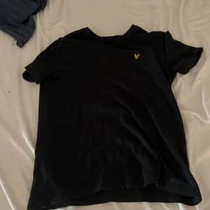 Svart t-shirt från Lyle & Scott - Svart t-shirt från Lyle & Scott med klassisk gul logga på bröstet. Enkel och stilren design med rund hals och korta ärmar. Perfekt för dig som gillar cleana och snygga plagg med diskret branding.