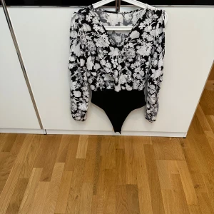 Svartvit blommig omlott-body - Snygg svartvit body med blommigt mönster och omlott-skärning framtill. Långa ärmar i lätt, flowigt tyg och v-ringad hals. Nedre delen är svart och åtsittande.