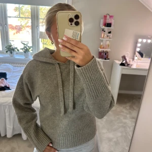 Grå stickad hoodie med huva - Mysig grå stickad hoodie med huva och snörning. Tröjan har breda ribbade muddar vid ärmslut och nederkant, samt en relaxed passform. Perfekt för kyliga dagar och enkel att styla med jeans eller mjukisbyxor.