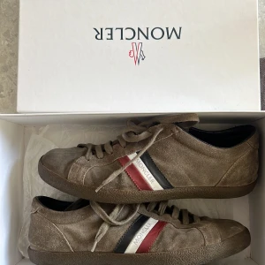 Bruna sneakers i mocka från Moncler Monaco ink box  - Snygga bruna sneakers från Moncler i mjuk mocka med klassiska Moncler-ränder i rött, vitt och svart på sidan. Skorna har rund tå, platt sula och snörning. Perfekta för dig som gillar stilrena och exklusiva sneakers med en sportig touch.