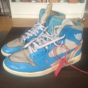 Nike Air Jordan 1 x Off-White UNC - Säljer ett par Nike Air Jordan 1 x Off-White UNC med ikoniska blå och vita paneler, klassisk Off-White-zip tie och textdetaljer på sidan. Skorna har högt skaft, vit snörning och coola orange accenter. Perfekt för dig som vill sticka ut med en streetwear-ikon. Väldigt använda genom åren därav priset. Från OGTony/TonySneaker