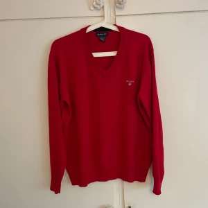 Röd v-ringad tröja från GANT - Klassisk röd v-ringad tröja från GANT med broderad logga på bröstet. Tröjan har långa ärmar, ribbade muddar och är tillverkad i 100% bomull. Perfekt för dig som gillar stilrena och tidlösa plagg med en poppig färg. Tröjan har storleken L, men passar bättre som en M.