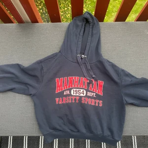 Mörkblå hoodie Manhattan H&M Divided - Mörkblå hoodie från H&M Divided med röd och vit text 'Manhattan Varsity Sports' på bröstet. Har huva med snörning och långa ärmar med röda och vita ränder. Skön och sportig stil, perfekt till jeans eller joggers.