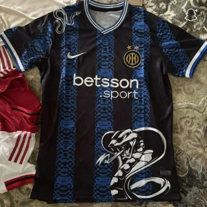 Inter Milan x Nike blå-svart fotbollströja - Snygg Inter Milan fotbollströja från Nike med blå och svarta ränder i ett unikt mönster. Stor vit orm tryckt på framsidan, klubbmärke med två stjärnor och vit Nike-logga. Rund hals med blå och vita detaljer. Materialet är lätt och andas, perfekt för match eller träning.