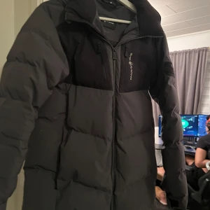 Svart dunjacka från Sail Racing - Säljer en svart dunjacka från Sail Racing med hög krage och huva. Jackan har två bröstfickor med dragkedja och logga på bröstet samt ärmen. Perfekt för kalla dagar och har en clean, sportig look med snygga detaljer.