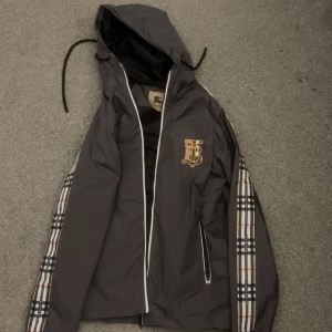 Grå Burberry vindjacka med huva - Snygg grå vindjacka från Burberry med klassiskt rutmönster längs ärmarna och stort guldigt logotryck på bröstet. Jackan har huva med snörning, dragkedja framtill och fickor med dragkedja. Perfekt för dig som vill ha en stilren och ikonisk look.