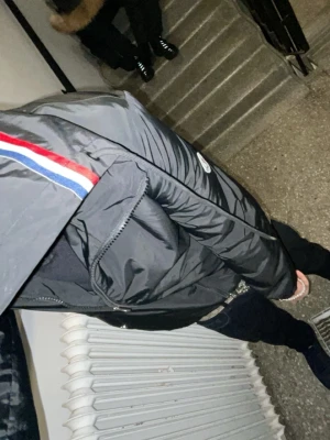 Svart Moncler vindjacka med huva - Svart vindjacka från Moncler med huva och dragkedja. Jackan har Moncler-logga på ärmen och en snygg röd, vit och blå rand på huvan. Materialet är polyester och jackan har en clean, sportig look med normal passform.
