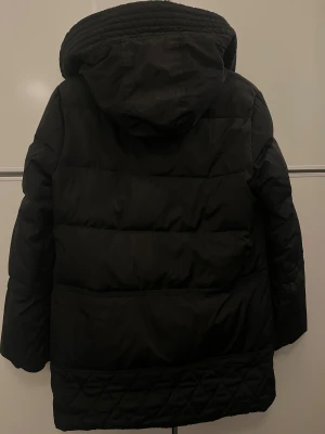 Svart pufferjacka med teddyfoder - Svart pufferjacka med quiltade detaljer längst ner och mysigt vitt teddyfoder på insidan. Jackan har huva, flera fickor med dragkedja och långa ärmar. Perfekt för kalla dagar när du vill hålla dig varm och ändå se chill ut. Står L på jackan men den passar M