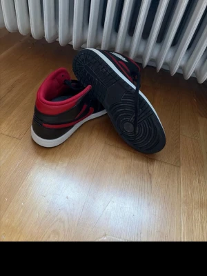Nike Air Jordan 1 svart/röd sneakers - Nike Air Jordan 1 mid sneakers i svart och rött med vit sula. Klassisk high-top modell med snörning och rund tå. Skorna har läderöverdel och ikonisk Air Jordan-logga på sidan. Röd swoosh och detaljer runt kragen ger en fet kontrast till den svarta basen.