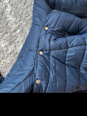 Blå quiltad pufferjacka från CRW - Säljer en blå pufferjacka från CRW med quiltad design och snygga guldfärgade knappar. Jackan har huva, dragkedja framtill och är tillverkad i polyester. Perfekt för dig som gillar stilren och funktionell ytterkläder.