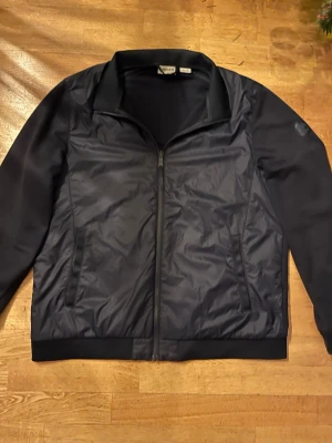 Svart vindjacka Timberland XXL - Svart vindjacka från Timberland i storlek XXL. Jackan har en stilren design med dragkedja framtill, två sidofickor och klassisk krage. Materialet är polyester och jackan har en normal passform. Perfekt för dig som gillar enkel och snygg streetstil.