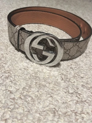 Gucci GG Supreme bälte med silverdetalj - Säljer ett Gucci GG Supreme bälte med klassiskt mönster och stor silverfärgad GG-logga som spänne. Bältet är beige och brunt med insida i ljusbrunt skinn. Perfekt för att lyfta din outfit med en lyxig touch.