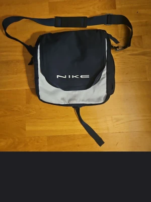 Nike axelväska i marinblå och grå - Säljer en stilren axelväska från Nike i marinblått och grått med orange detaljer på insidan. Väskan har justerbar axelrem med vadderad del, flera fack med dragkedja och tydlig Nike-logga på framsidan. Perfekt för att bära med sig laptop, böcker eller andra prylar.