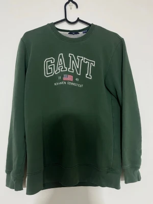 Gant Tröja (Grön) - Mörkgrön sweatshirt från GANT med klassisk logga och amerikansk flagga broderad framtill. Tröjan har rund hals, långa ärmar och ribbade muddar. Perfekt för en chill och stilren look.